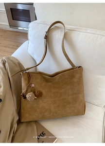 Detail of a brown suede tote bag with contrast stitching and pom-pom charm on a sofa