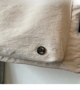 Detailed view of beige crinkled fabric and circular CM metal badge on the tote bag