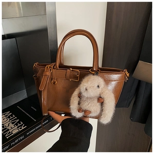 Close-up of brown glossy leather tote bag featuring decorative straps and a plush bunny charm