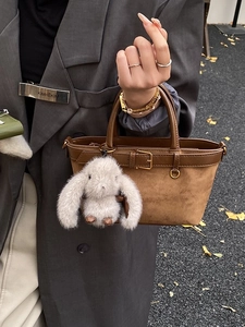 Brown suede bucket bag with leather handles and a plush rabbit charm in an outdoor setting