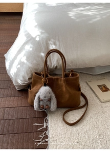Brown suede tote bag with gray faux fur charm and long strap in a cozy home setting