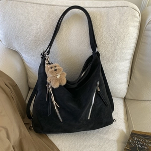Detail of a black matte tote bag with dual front zippers and a plush bear charm on a sofa