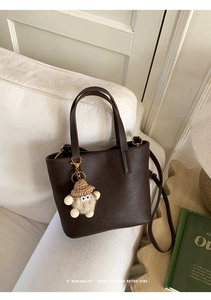 Detail of dark brown lychee leather bucket bag with cute plush doll charm on white sofa