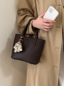 Minimalist brown leather bucket bag with plush charm shown with casual trench coat