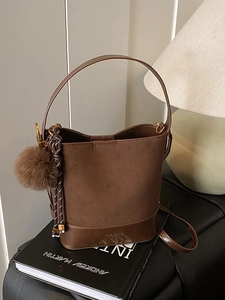 Brown suede bucket bag with leather trim and pompom detail on a minimalist background