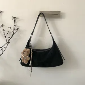 Detail of black suede crescent bag featuring a brown teddy bear pendant on white wall