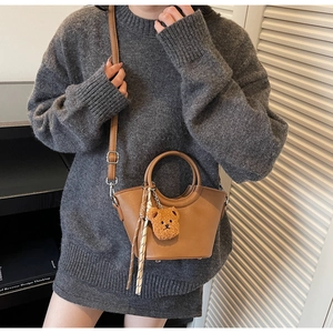Dettaglio di una borsa a secchiello in pelle marrone con ciondolo a orsetto e maglione grigio