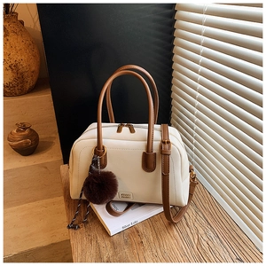 Detail of cream Boston bag with brown handles, plush charm, and adjustable strap on a desk