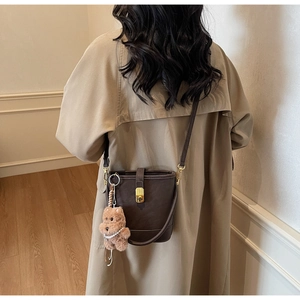Détail du sac seau marron foncé avec fermoir doré et breloque ours en peluche sur trench