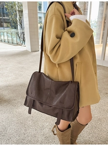 Detail view of a large brown messenger bag worn over a beige coat in a soft lit hallway