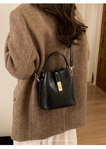 Détail d'un sac seau en cuir noir à fermoir doré porté sur un manteau en laine brun
