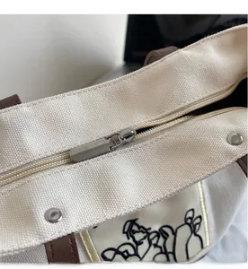 Detail view of silver metal zipper and brown leather handles on off-white canvas tote bag