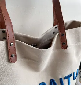 Detail of canvas tote bag showing blue embroidery, leather handle and rivet accents