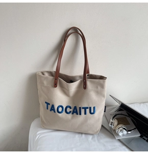 Detail of canvas tote bag featuring blue embroidered letters and brown leather handles