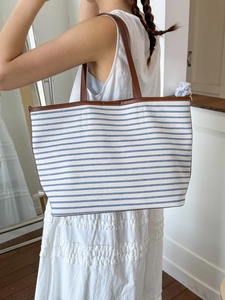 Large striped canvas tote bag with brown leather handles worn by a model in white dress