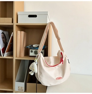 Detail of cream nylon crescent crossbody bag with red logo and plush charm on wooden shelf