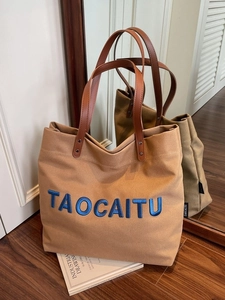 Brown canvas tote bag with leather straps and blue embroidery on a warm wooden floor
