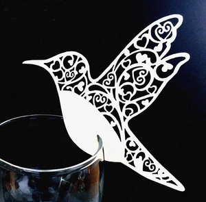 Laser-cut white hummingbird paper place card with heart patterns on a wine glass rim