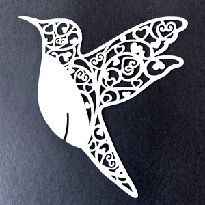 Laser-cut white hummingbird paper place card with intricate floral heart pattern on black background