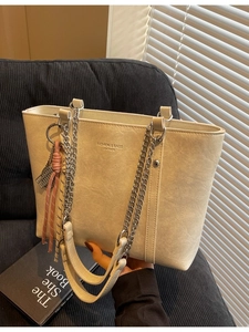 Beige faux leather tote bag with silver chain straps and tassel detail on a sofa