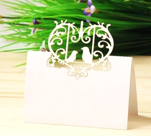 Laser-cut white wedding place card with intricate bird and heart motifs on wood table