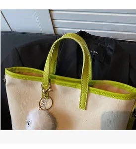 Close-up of a canvas tote bag featuring green leather trim and a plush ball charm detail