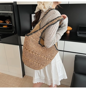 Detail of a large woven straw shoulder bag with black trim worn by a model in a kitchen