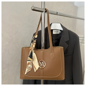Detail of brown tote bag with white stitching, metallic M logo and decorative silk scarf