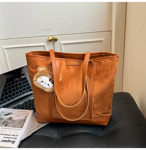 Detail of caramel suede tote bag with brown plush bear charm on a black leather bench