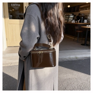 Detail of a brown patent leather box bag with gold zipper held by a model in a gray coat
