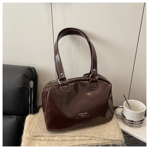 Detail einer dunkelbraunen Hochglanz-Boston-Bag mit Metall-Logo auf beigem Schal