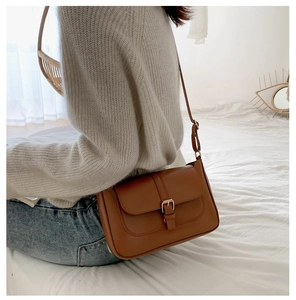 Brown leather crossbody bag with golden buckle shown on a model in a minimalist home setting