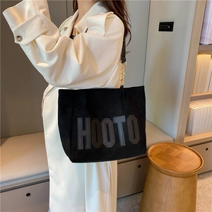 Large black suede tote bag with HOOTO lettering and gold chain worn with trench coat