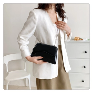 Detail of a black patent leather crossbody bag with slim metal chain on a female model