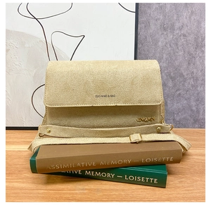 Detail der beige Wildleder-Umhängetasche mit Markenlogo und Metallbeschlägen auf Holztisch