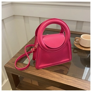 Close-up of a magenta trapezoid crossbody bag with round handle on a glass table