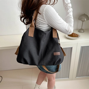 Large black canvas tote bag with brown straps shown on shoulder in minimalist setting