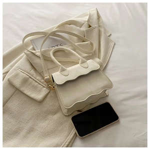 Detail of cream white mini crossbody bag with wavy flap design next to a smartphone