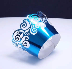 Detailed laser-cut blue cupcake wrapper with intricate hollow scroll patterns on white surface