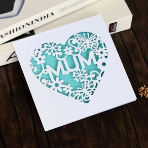 Close-up of white laser-cut card with MUM text and butterfly heart pattern on wood