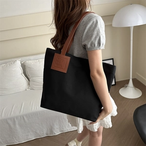 Large black canvas tote bag with brown leather handles worn by a model in minimalist home setting