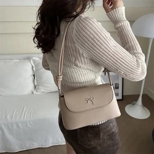 Elegant beige crossbody flap bag with silver bow accent in a minimalist indoor setting