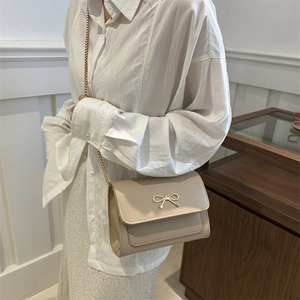 Sac bandoulière beige en similicuir avec nœud doré et chaîne, présenté sur un modèle