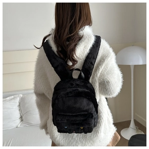 Model wearing a black plush backpack with colorful button eyes in a cozy room setting