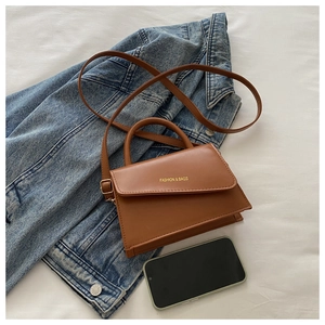 Brown leather handbag with FASHION & BAGS gold text next to denim jacket and phone