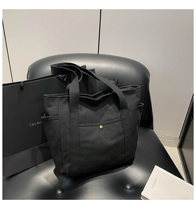 Detail view of black canvas tote bag featuring a metal snap button pocket and reinforced straps
