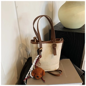 Detail of beige bucket bag with brown trim, featuring a teddy bear charm and fashion text