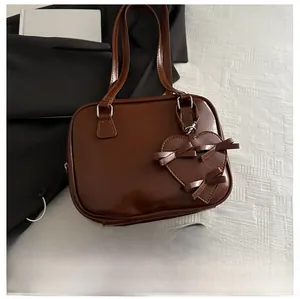 Brown faux leather handbag featuring a large bow and heart charm on a white fabric surface