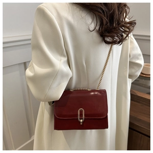 Model wearing a burgundy patent leather crossbody bag with gold chain and oval buckle