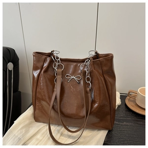 Detail of a brown glossy tote bag featuring a silver metal bow and chunky chain strap